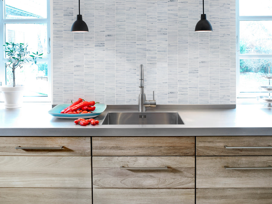 Modern kitchen with light wood cabinets, stainless steel sink, and a Luna Kit Kat Glass Tile backsplash. Two black pendant lights hang above a counter with red peppers, while large windows fill the space with natural light.