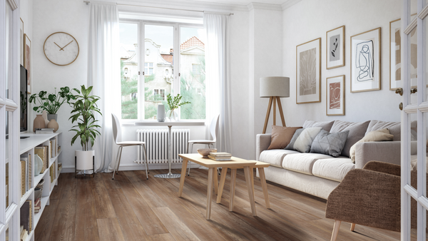 A bright, modern living room with wooden floors, a beige sofa, a wooden coffee table, a brown armchair, wall art, indoor plants, a floor lamp, and large windows with white curtains letting in natural light.