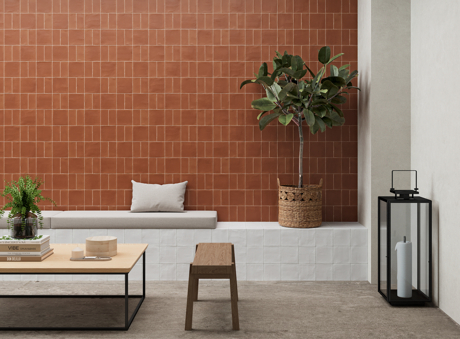 A modern minimalist living space features a red Harmony 5x5 Porcelain Tile accent wall, built-in white bench with cushion, potted plant, wooden stool, coffee table with books and decor, and a large lantern with a white candle.