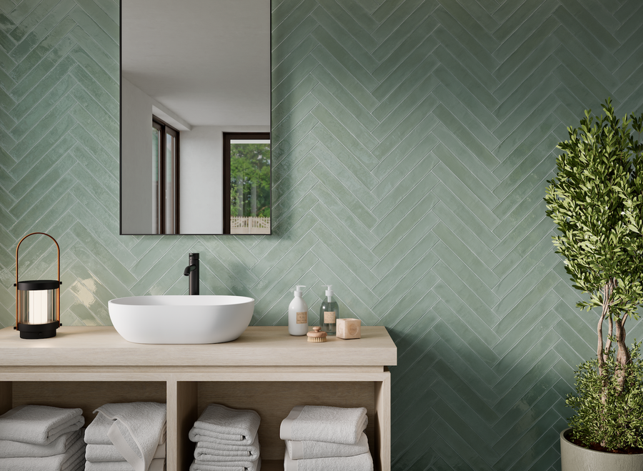 Modern bathroom with a green herringbone tile wall, rectangular mirror, white vessel sink, black faucet, and wooden vanity with neatly folded towels, toiletries, a lantern, and a potted plant.
