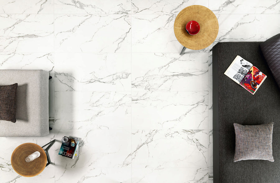 A top-down view of a marble-tiled floor with a gray sofa, a beige armchair, two round wooden side tables, a red cup, a magazine, and decorative pillows arranged neatly.