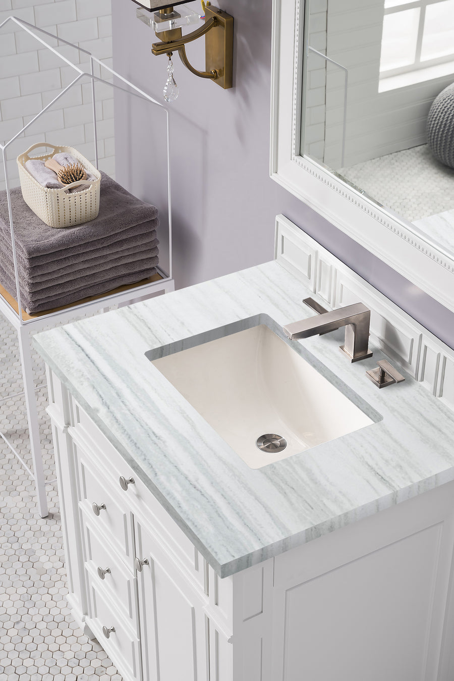 A modern bathroom sink with a rectangular basin, marble countertop, and stainless steel faucet. A stack of folded gray towels and a basket with toiletries sit on a nearby shelf. White cabinetry and a large mirror are visible.