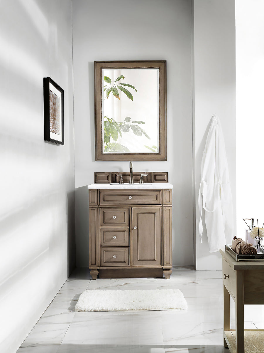A modern bathroom showcases the Bristol 30" Vanity, complemented by a large framed mirror reflecting leafy branches, a white rug on marble floors, a robe on the wall, and minimalist décor for a bright, stylish space.