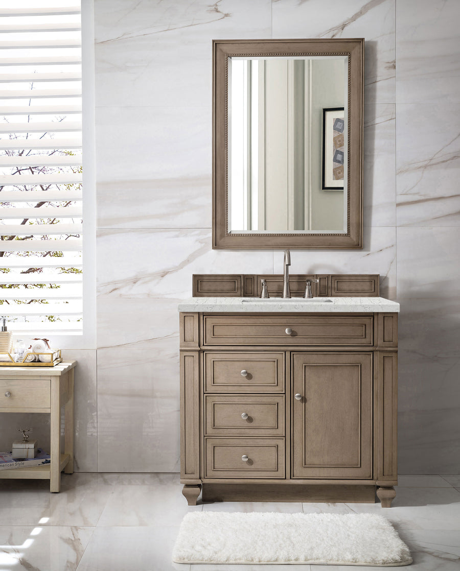 A bathroom featuring the Bristol 36" Vanity, a marble countertop, framed mirror, sleek drawer handles, a white rug, side table, and sunlit white marble walls showcases elegant transitional design.