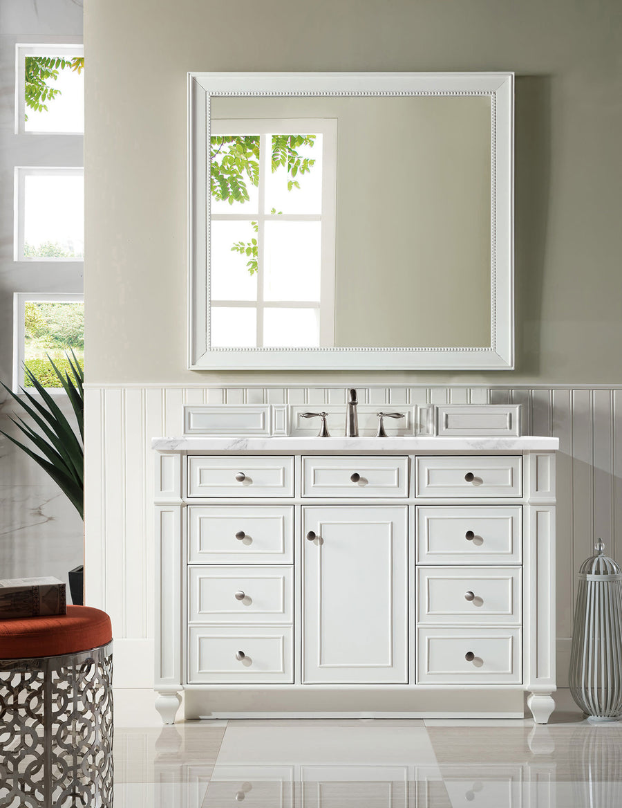 A bright bathroom showcases the Bristol 48" Vanity with ample storage and bronze hardware, topped with a sink. A large framed mirror above reflects greenery, while beige walls and white wainscoting complete the look.