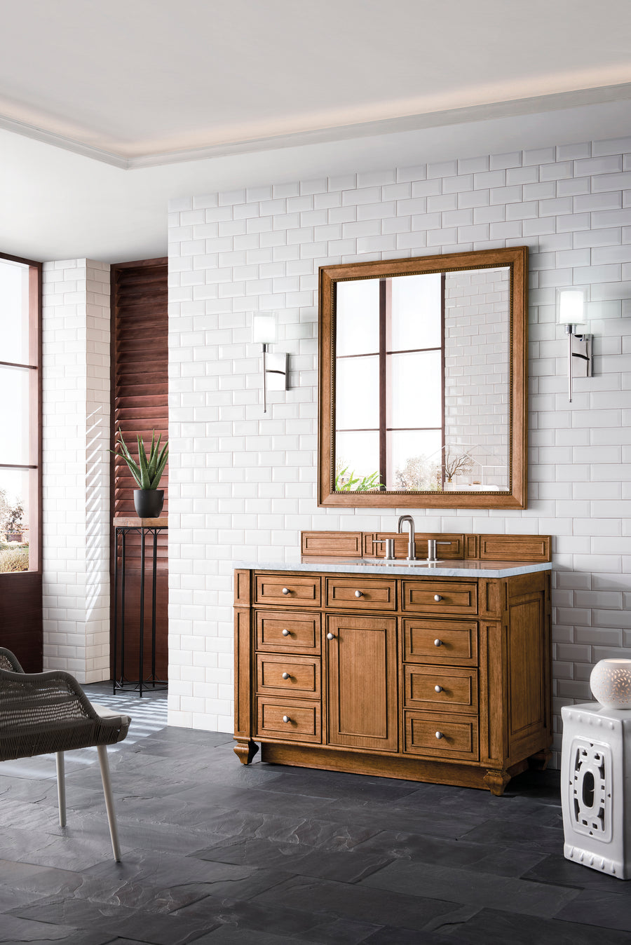 A bright bathroom with white subway tile walls, a large wooden vanity with drawers, a matching framed mirror, two wall sconces, a potted plant, a modern chair, and slate tile flooring.