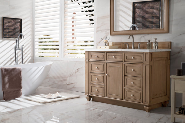 A bright, modern bathroom with a freestanding white bathtub, wooden vanity with drawers and a countertop sink, large mirror, towel rack, and sunlight streaming through white slatted blinds.