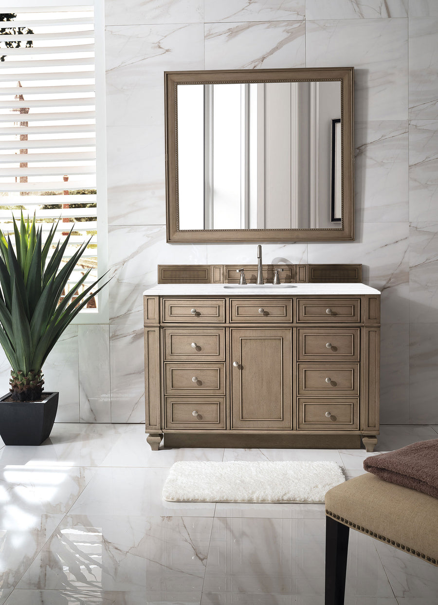 A modern bathroom showcases the Bristol 48" Vanity with a rectangular mirror, white countertop, silver fixtures, potted plant, cream rug, and beige bench on glossy marble as sunlight streams through window blinds.