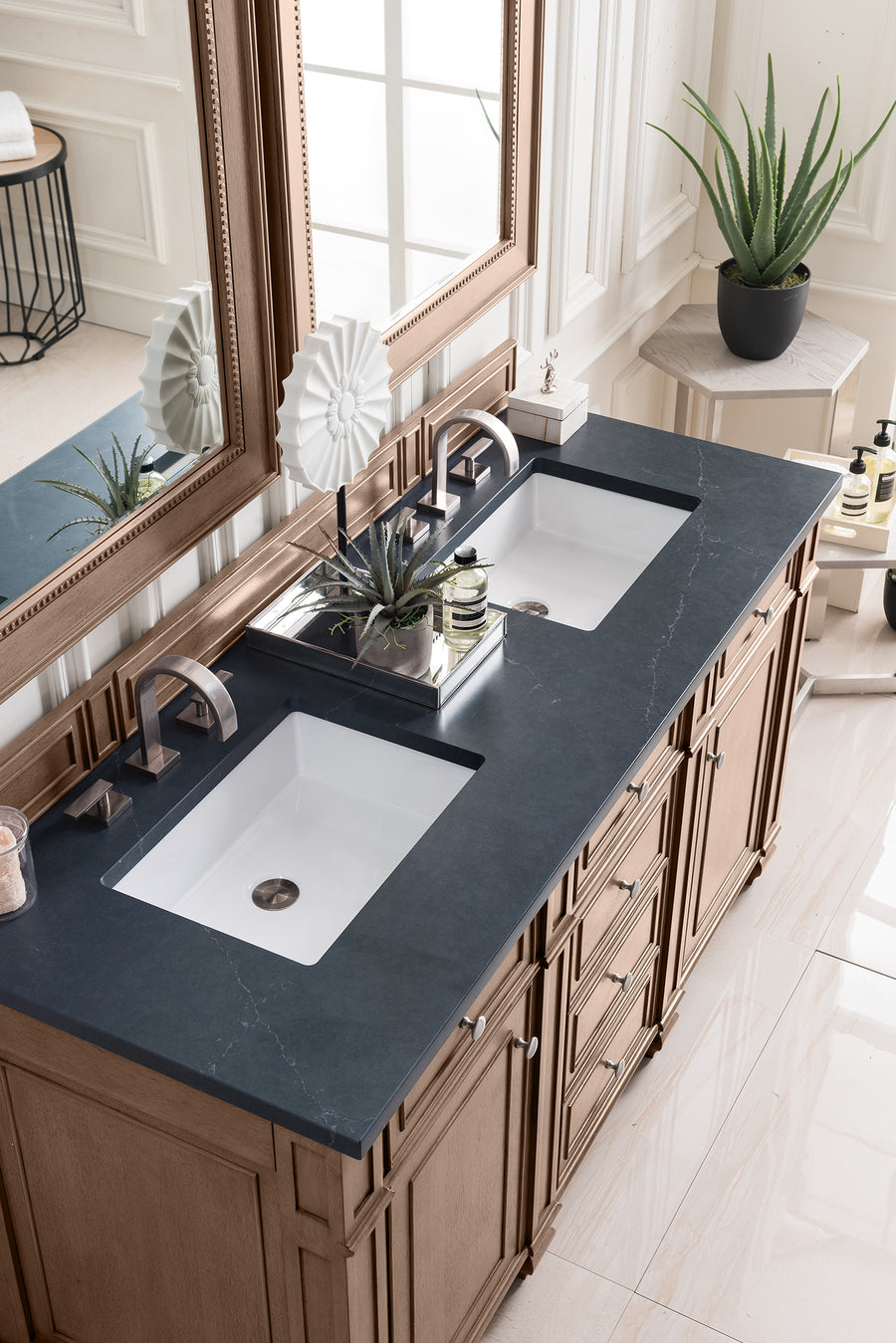 Double bathroom vanity with a black countertop, two white sinks, bronze fixtures, a large mirror, potted plants, and toiletries arranged neatly; natural light streams in from a nearby window.