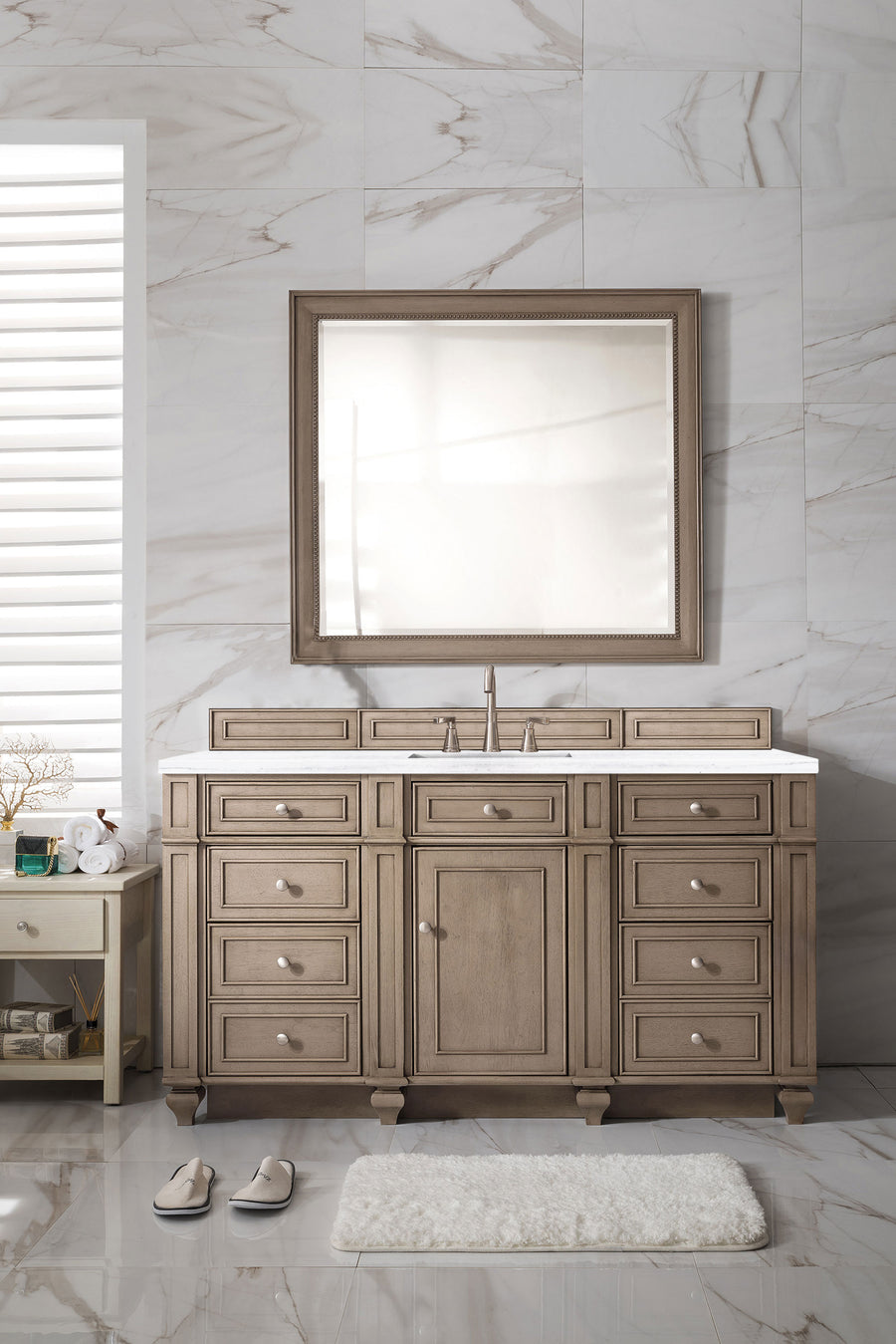 A luxury bathroom with beige marble walls, a large mirror above a Bristol 60" Vanity with multiple drawers, plus a white rug, slippers, and a side table for towels and toiletries.