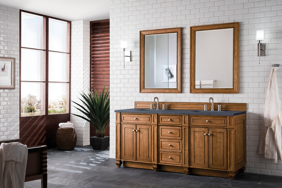 A modern bathroom with white subway tile walls, a wooden double-sink vanity, two mirrors, wall sconces, a large plant, a towel on a hook, and natural light coming from large windows.