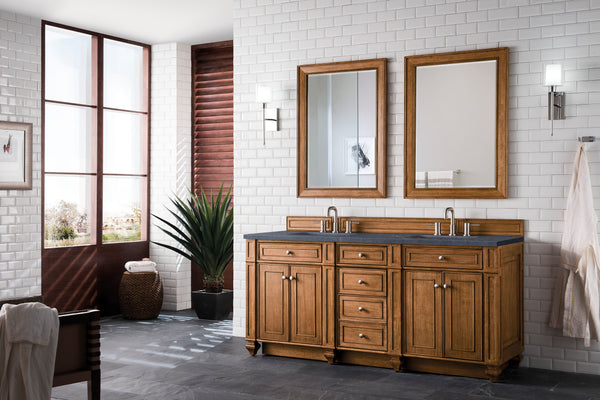 A modern bathroom with white subway tile walls, a wooden double-sink vanity, two mirrors, wall sconces, a large plant, a towel on a hook, and natural light coming from large windows.