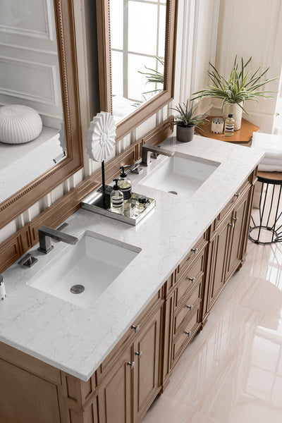 A modern bathroom with a double sink vanity, marble countertop, two rectangular mirrors, potted plants, toiletries on a tray, and wooden cabinets below. Natural light streams in from a tall window.