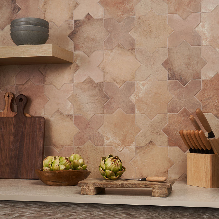 A kitchen counter with wooden cutting boards, a bowl of artichokes, a knife block, and stacked bowls. The backsplash features tan and beige star-patterned tiles.