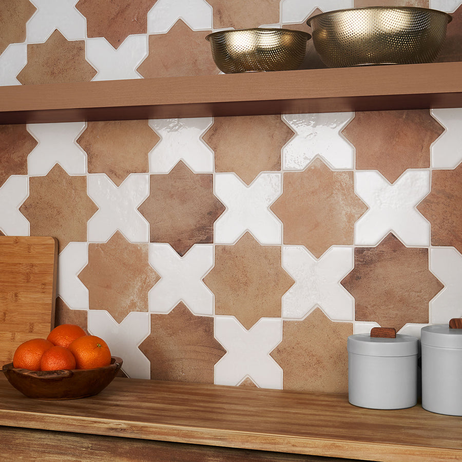 A kitchen counter with a wooden bowl of oranges, two white canisters, a cutting board, and a shelf holding two textured metal bowls. The backsplash features geometric star-shaped tiles in brown and white tones.