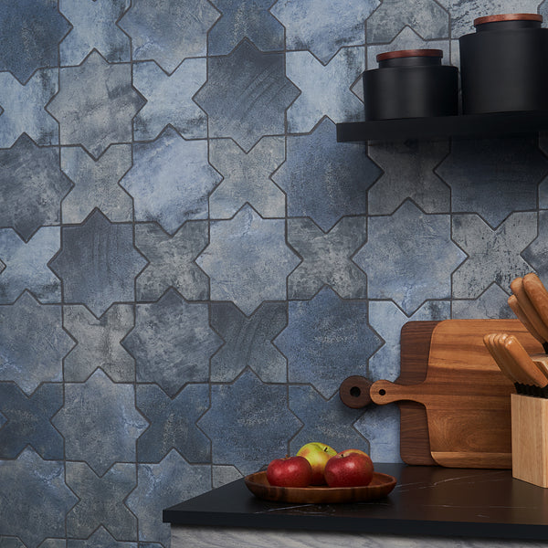 A kitchen countertop with apples, wooden cutting boards, and knives in a holder sits in front of a wall with blue star-shaped patterned tiles. Black jars with lids are on a shelf above.
