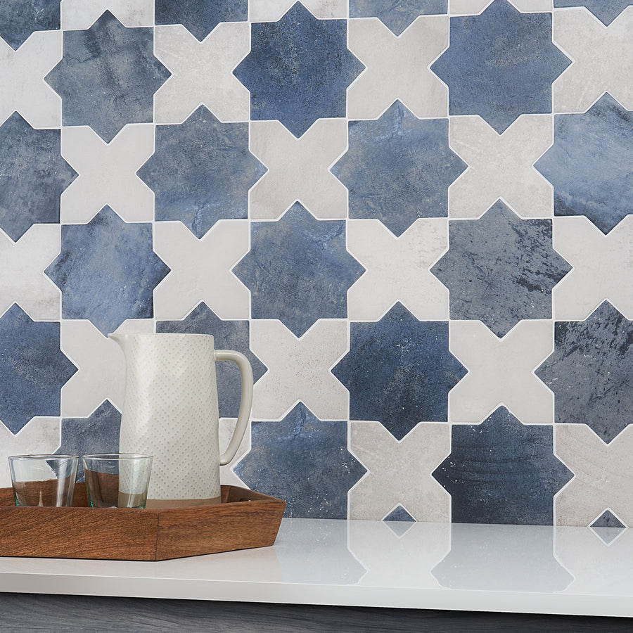 A decorative wall with blue and white star-shaped tiles is shown behind a wooden tray holding a white textured pitcher and two drinking glasses on a white countertop.