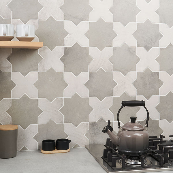 A modern kitchen features a patterned tile backsplash, a stovetop with a gray kettle, black condiment containers, a gray cup, and two glasses on a wooden shelf. The decor is minimalist with neutral tones.
