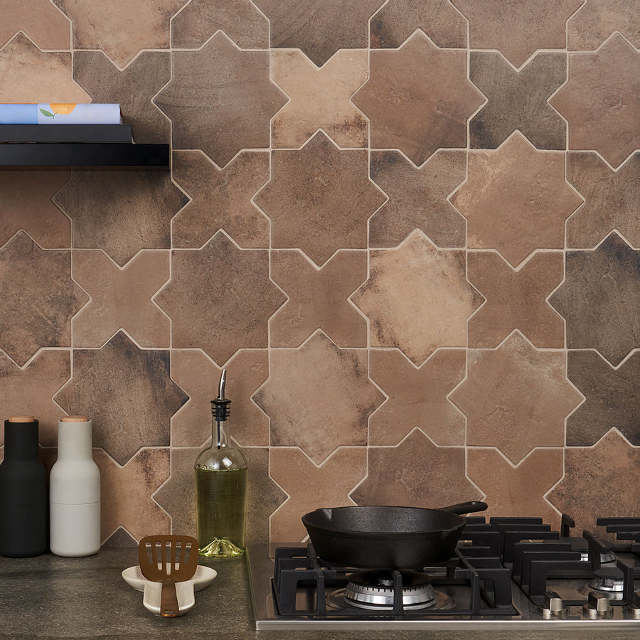 A kitchen countertop with a gas stove, olive oil bottle, utensils, and containers. The backsplash features interlocking star-shaped brown and beige tiles with varied earthy tones. A black shelf holds a book and small item above.