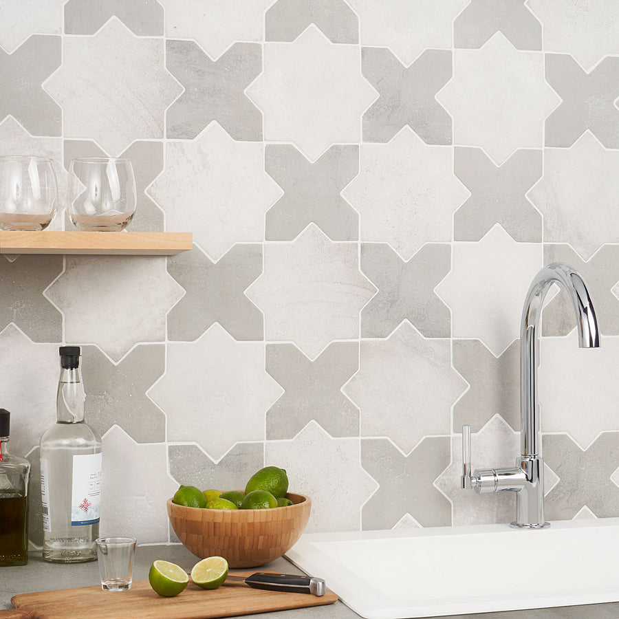 A modern kitchen scene with a geometric tile backsplash, a chrome faucet, a wooden bowl filled with limes, a wooden shelf holding glasses, and bottles on the countertop next to a sink.