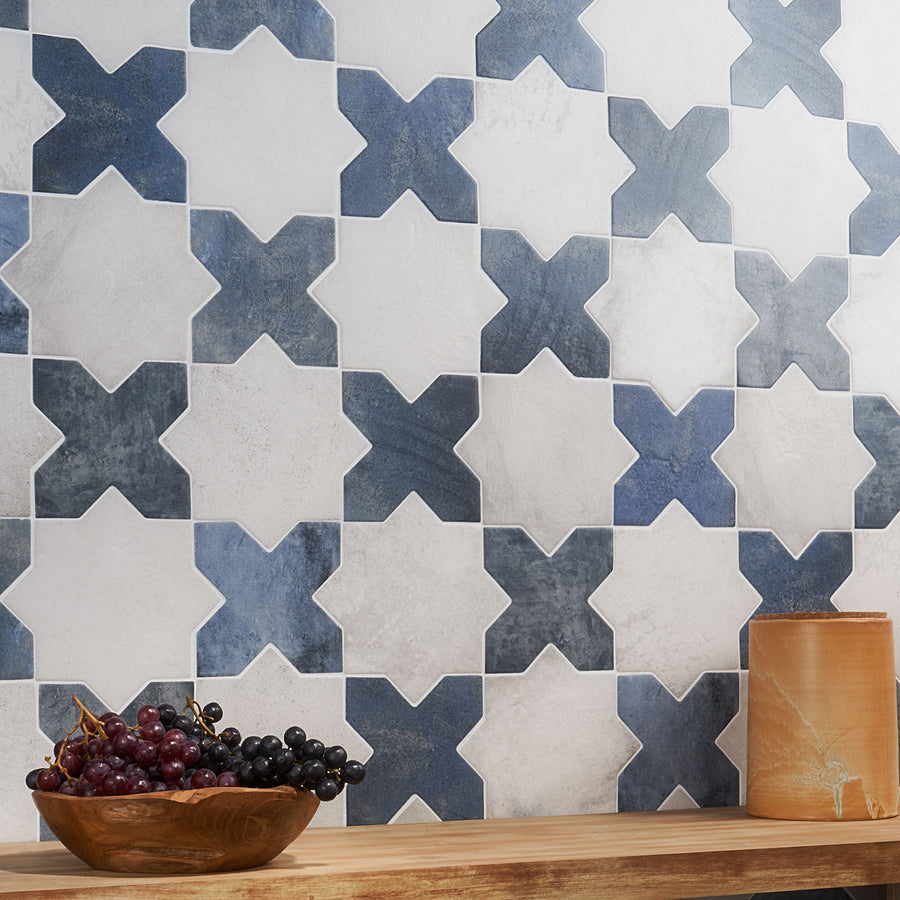 A wall with a geometric pattern of blue and white X-shaped tiles, a wooden shelf below, a bunch of grapes, and a brown ceramic vase on the right side of the shelf.