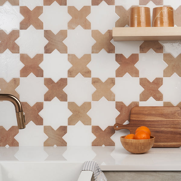 A kitchen backsplash with a geometric tile pattern of tan and white X-shapes, a copper faucet, a wooden cutting board, a bowl of oranges, and wooden shelves holding brown cups.