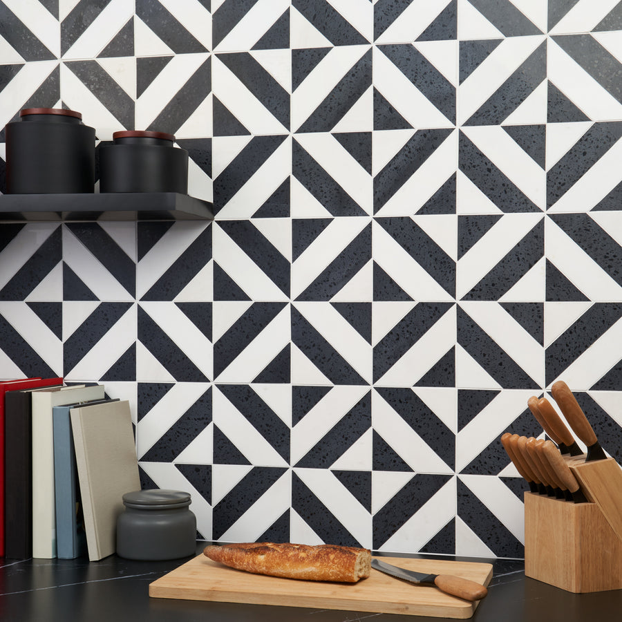 A modern kitchen with a black and white geometric tiled backsplash, black shelves with jars, a stack of books, a breadboard with a baguette and knife, and a wooden knife block on a dark countertop.