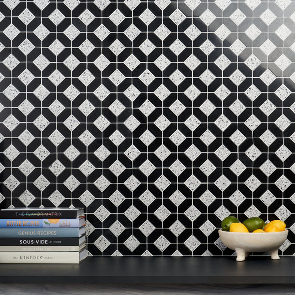 A patterned black and white tile wall with geometric shapes, a dark countertop, a stack of cookbooks on the left, and a white bowl filled with lemons and limes on the right.