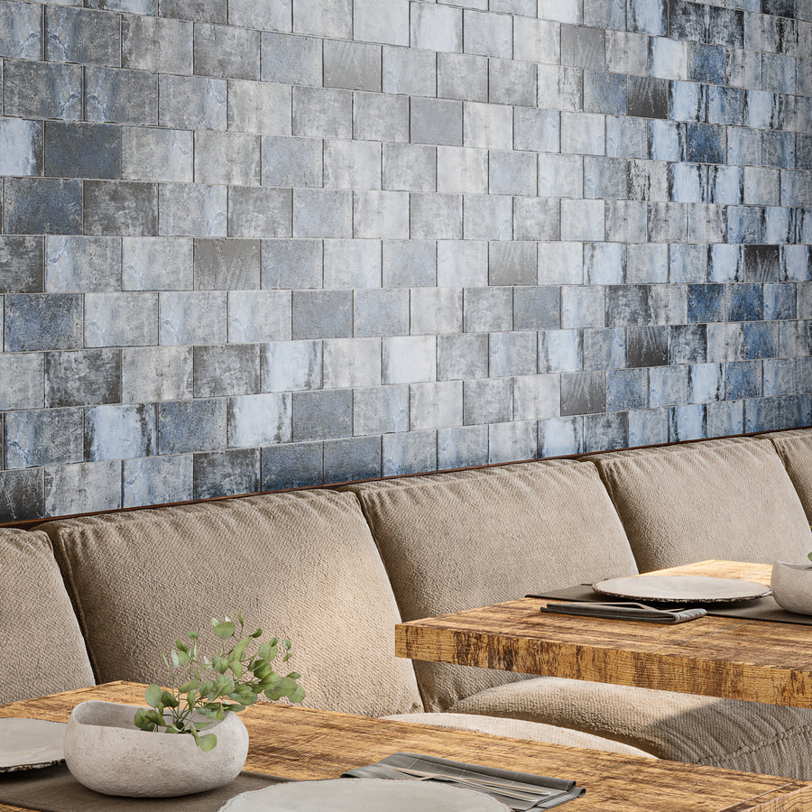 A cozy dining area with a beige upholstered bench, rustic wooden tables set with plates, and a decorative plant. The wall features a textured, blue-gray tile pattern, adding a modern touch to the space.