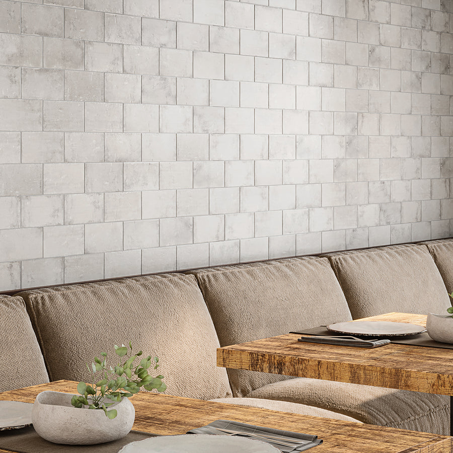 A cozy dining area with a beige cushioned bench, wooden tables, simple table settings, and small potted plants. The wall is covered with light gray, textured subway tiles arranged in a grid pattern.