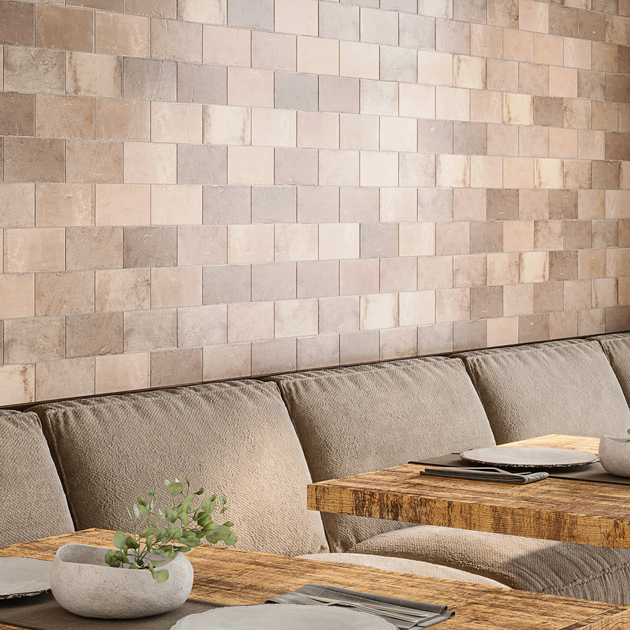 A cozy seating area with beige upholstered benches, rustic wooden tables set with plates and cloth napkins, and a wall covered in square, earth-toned tiles. A small stone vase with greenery sits on one table.