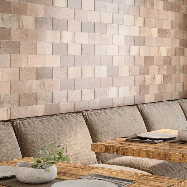 A cozy seating area with beige upholstered benches, rustic wooden tables set with plates and cloth napkins, and a wall covered in square, earth-toned tiles. A small stone vase with greenery sits on one table.