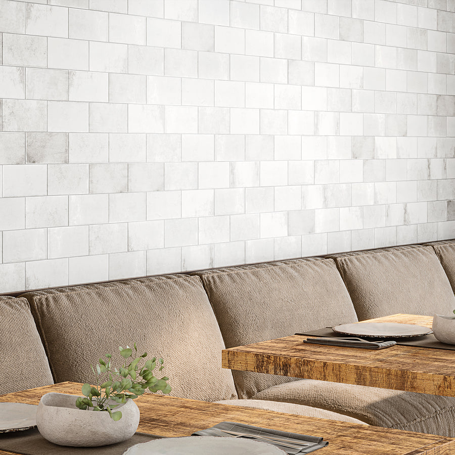 A cozy dining area with beige cushioned seating, rustic wooden tables set with plates and napkins, a small plant centerpiece, and a white subway tile wall in the background.
