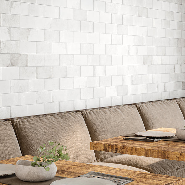 A cozy dining area with beige cushioned seating, rustic wooden tables set with plates and napkins, a small plant centerpiece, and a white subway tile wall in the background.