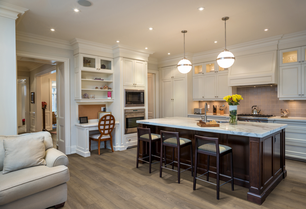 Modern kitchen with white cabinets, marble island with dark wood base, three bar stools, pendant lights, stainless steel appliances, a built-in desk with a chair, and a vase of yellow flowers on the counter.