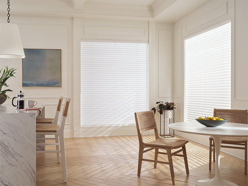 A bright, modern dining area features large windows dressed with Aria™ Soft Blinds, wooden chairs, a round table topped with lemons, and a marble countertop. Sunlight pours in, illuminating the light wood floors.