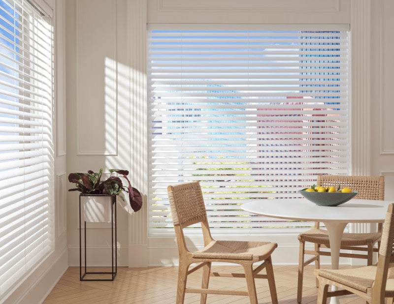 A bright dining area features large windows fitted with cordless Aria™ Soft Blinds, allowing sunlight in. Three woven chairs circle a round white table topped with a bowl of lemons. A potted plant adds greenery near the window’s cityscape view.