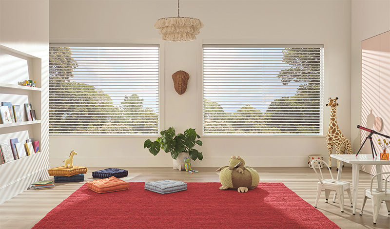 Bright, airy kids’ playroom with two large windows featuring Aria™ Soft Blinds, a red rug, cushions, plush animal toys, a giraffe figurine, bookshelves on the left, a white desk and chair, and indoor plants by the window.