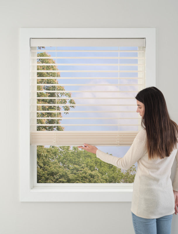 A woman with long dark hair and a light sweater adjusts Aria™ Soft Blinds by a window, enjoying gentle light control as green trees and a blue sky with clouds appear outside.
