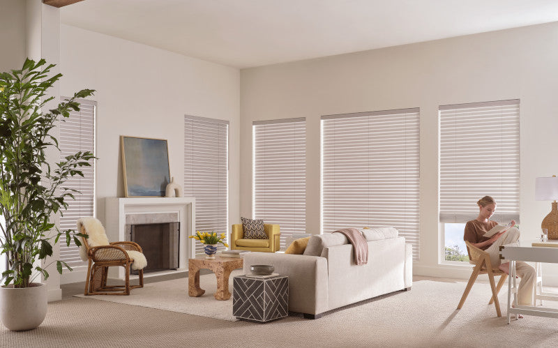 A bright living room with five large windows fitted with Aria™ Soft Blinds, neutral furniture, a potted plant, and a person reading at a desk. The space also includes a sofa, armchair, fireplace, and decorative accents.