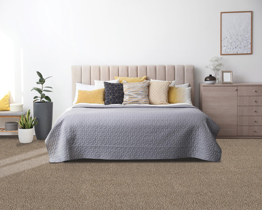 A modern bedroom features a beige upholstered bed with a gray quilt, several decorative pillows in yellow, white, and gray, a wooden nightstand with decor, potted plants, and a framed wall art on a white wall.