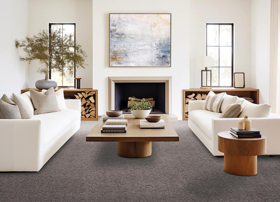 A modern living room with two white sofas facing each other, a wooden coffee table in the center, a fireplace with stacked firewood on both sides, large windows, and abstract art above the mantel.