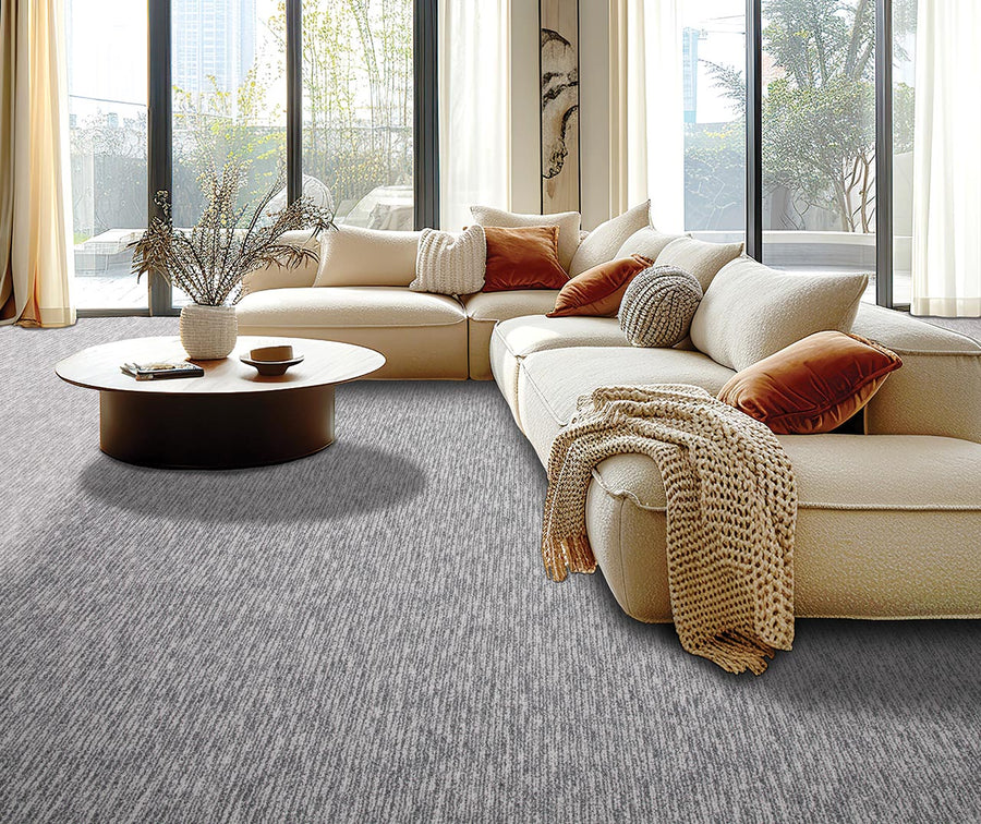 A modern living room with a large cream sectional sofa, assorted beige and rust-colored pillows, a round wooden coffee table with vases, and floor-to-ceiling windows letting in natural light. Gray carpet covers the floor.