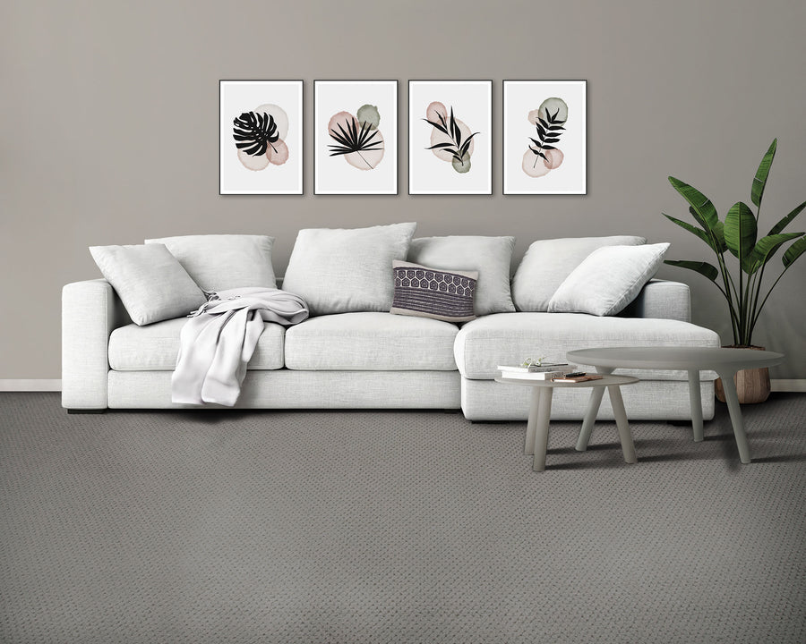 A modern living room with a light gray sofa, patterned pillow, and a throw blanket, three minimalist botanical prints on the wall, a round coffee table, and a tall potted plant beside the sofa.