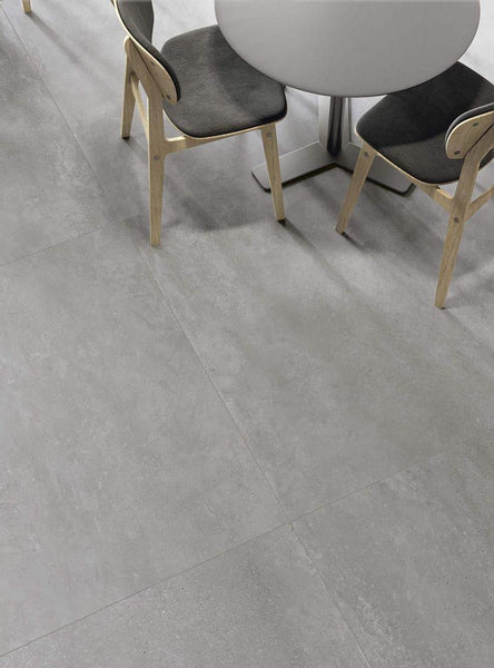 A round light gray table with two light wood chairs featuring dark gray seat cushions sits on a large, smooth, light gray tiled floor. The scene has a modern and minimalist aesthetic.