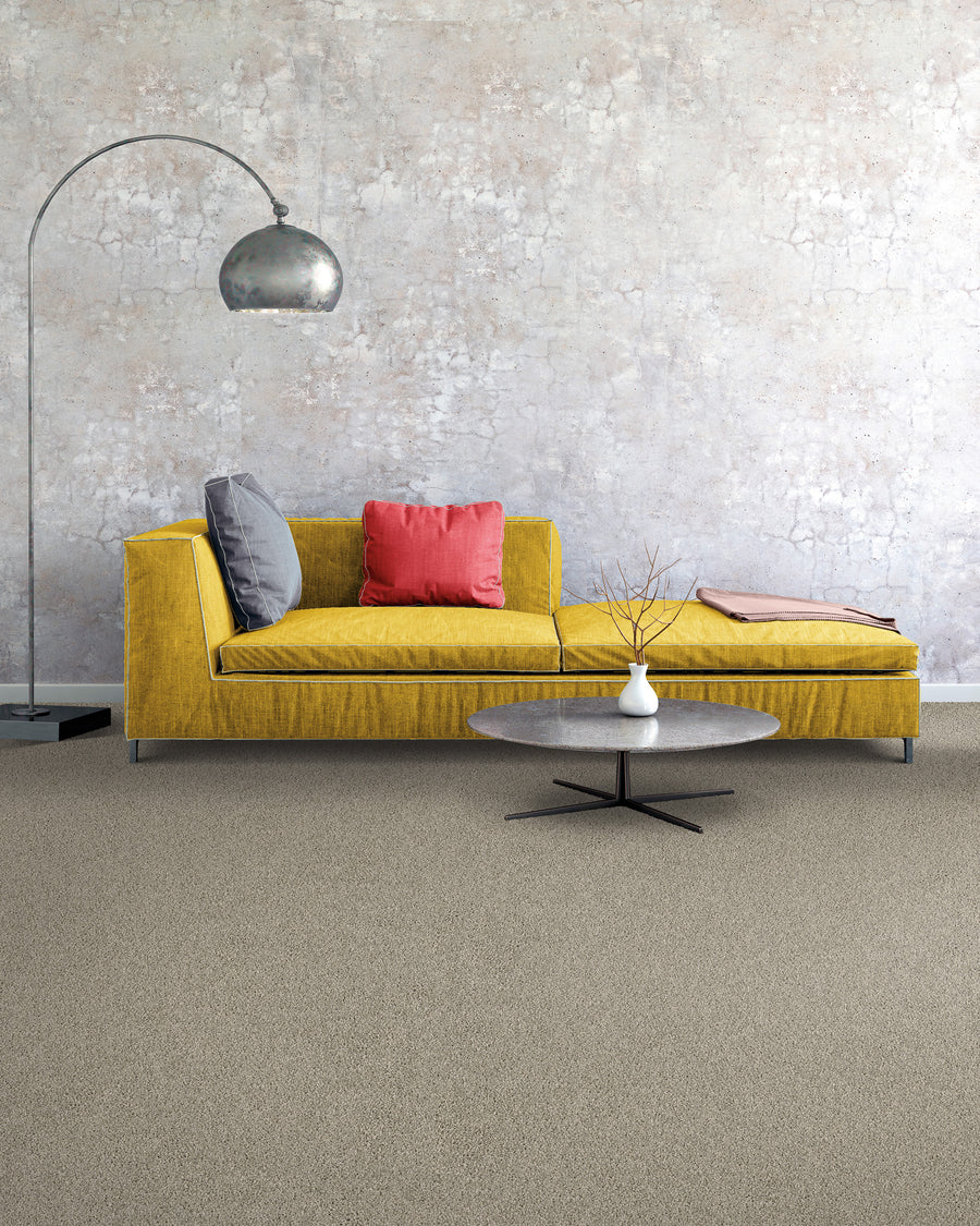 A modern living room with a yellow sofa, pink and gray cushions, a round coffee table, a floor lamp, and a textured, light-colored wall background. The floor is carpeted in a neutral tone.