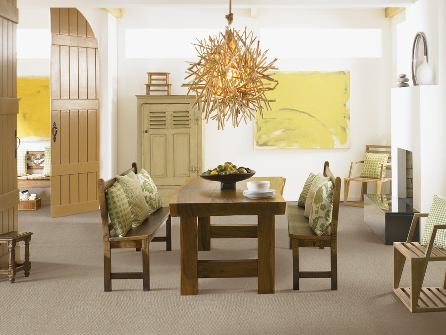 A modern dining room with a wooden table, benches with green-patterned pillows, a wooden chandelier, a yellow abstract painting, neutral walls, and natural light from high windows.