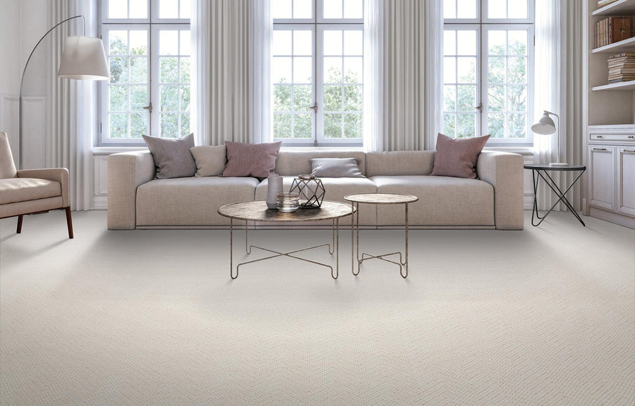 A modern living room with a beige sofa, pastel throw pillows, two round coffee tables, large windows with sheer curtains, a floor lamp, and built-in shelves on the right. Natural light fills the space.