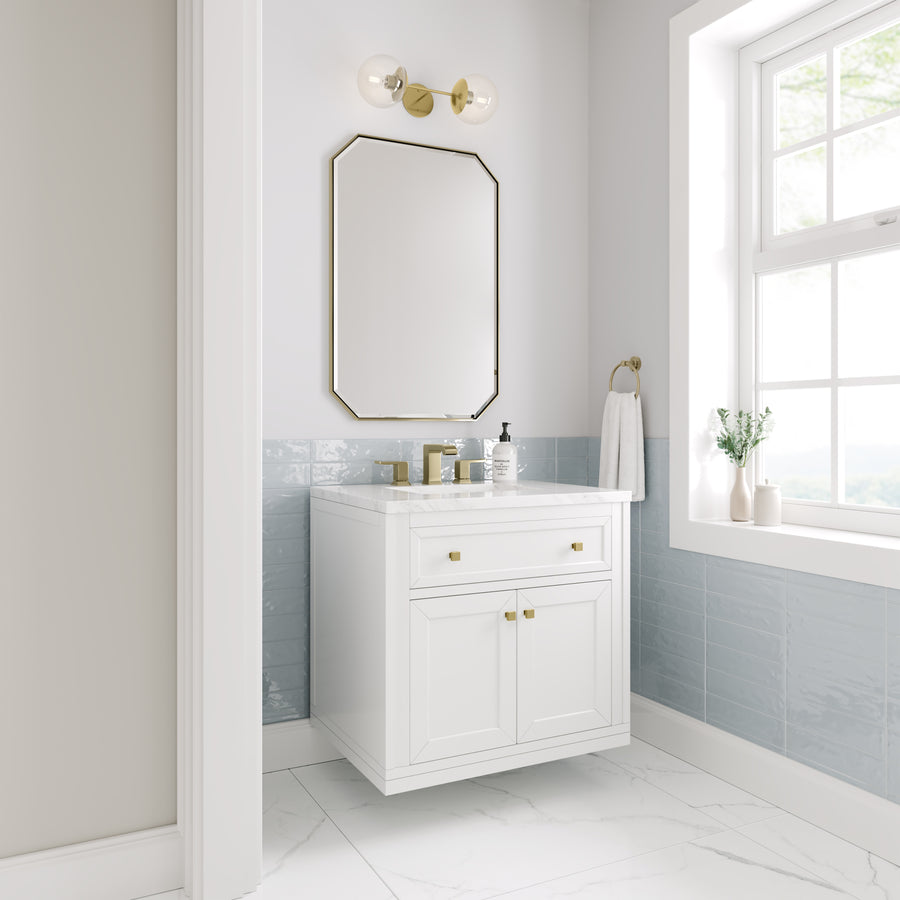 Bright bathroom with a white vanity, gold hardware, octagonal mirror, wall sconce, light blue tile backsplash, marble floor, towel ring, and a window letting in natural light. A plant and soap dispensers sit on the counter.