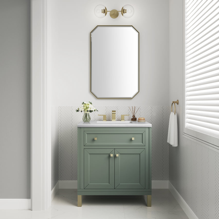 A modern bathroom features a Chicago 30" Vanity in green with gold hardware, a gold-trim rectangular mirror, flowers in a vase, and a towel on the wall. Sunlight enters through white blinds covering the window.